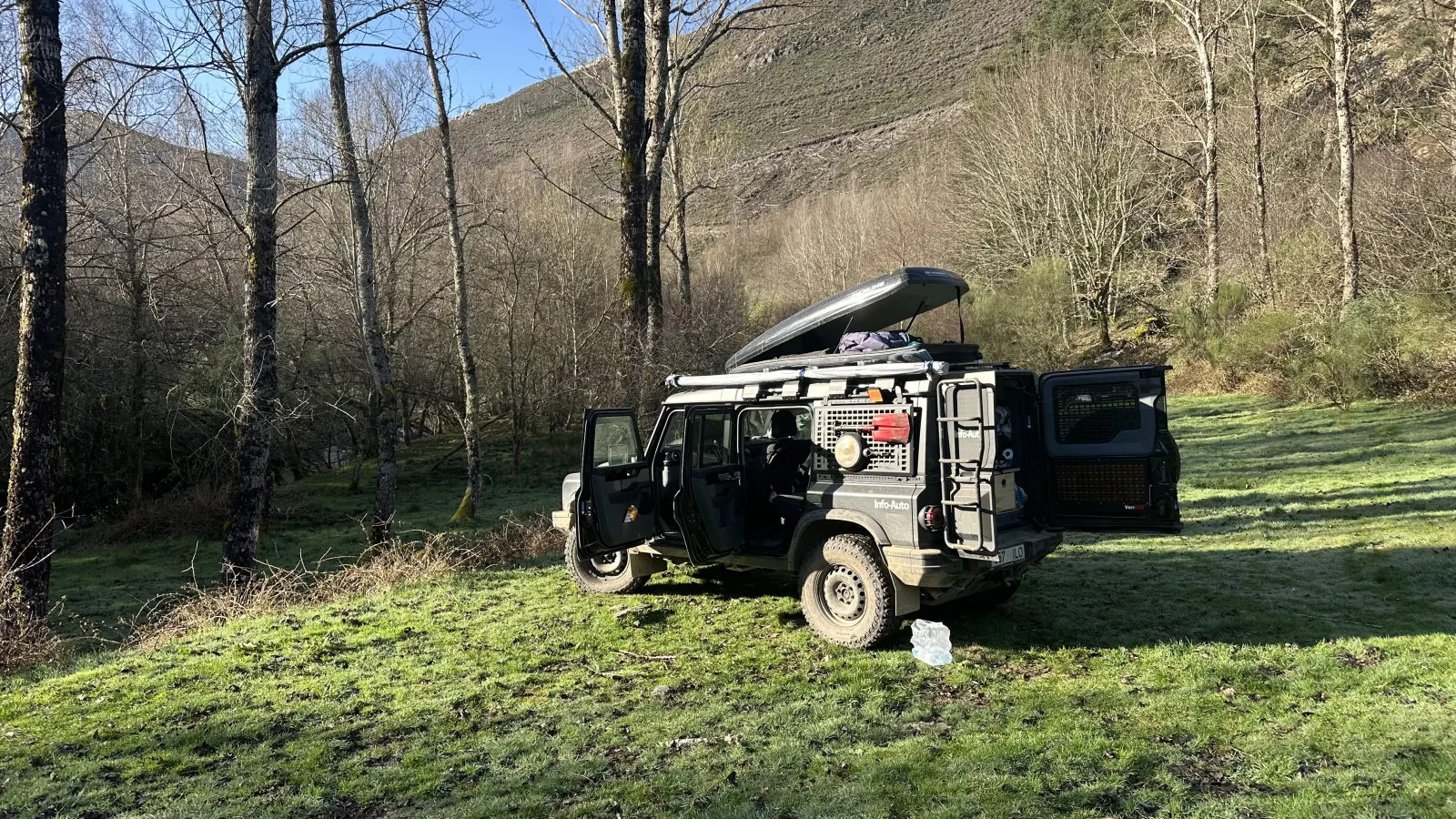 A dark expedition 4x4 with its doors and roof box open, parked on a frosty meadow among bare trees below a mountain slope